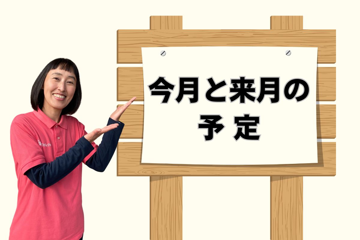 成田あすかが笑顔で立て看板を指し示しながら、「今月と来月の予定」を説明しています。