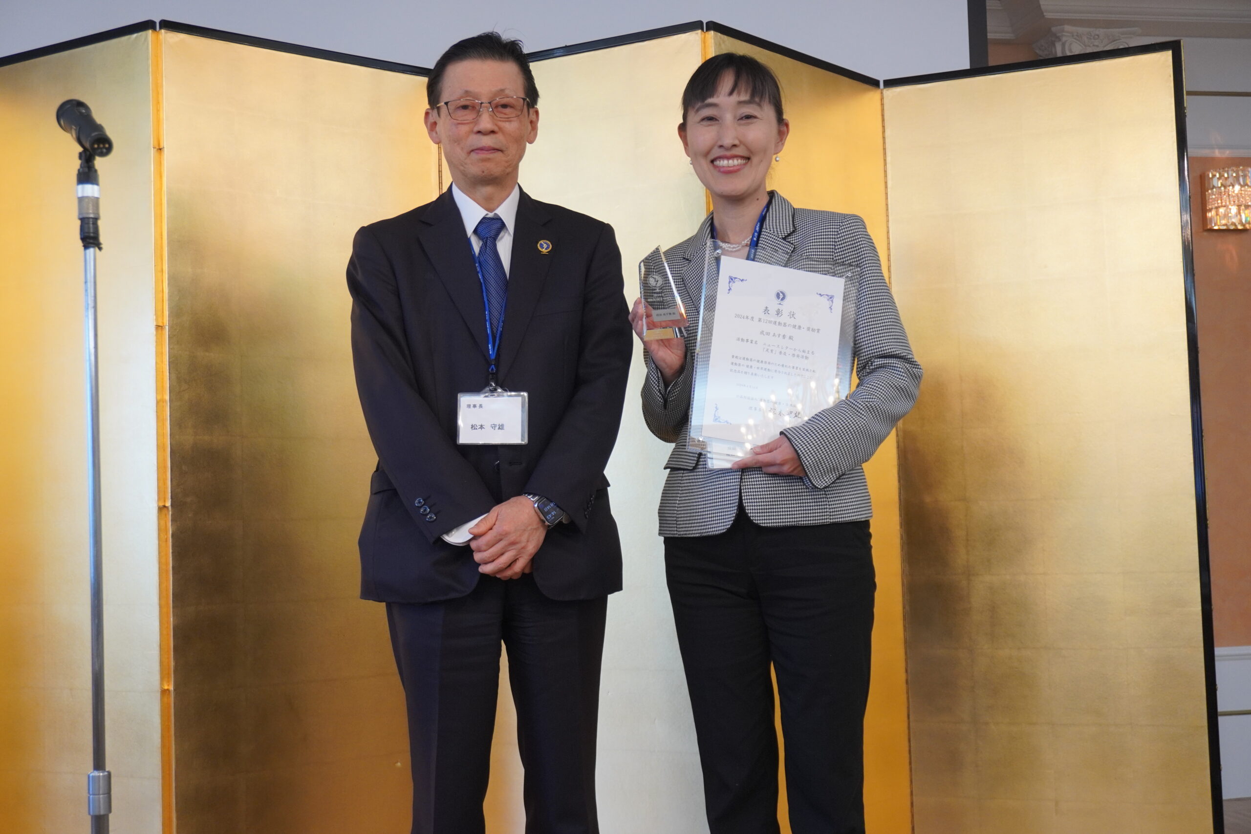 運動器の健康・日本協会の理事長である松本守雄様と、表彰状と盾を手に笑顔の成田