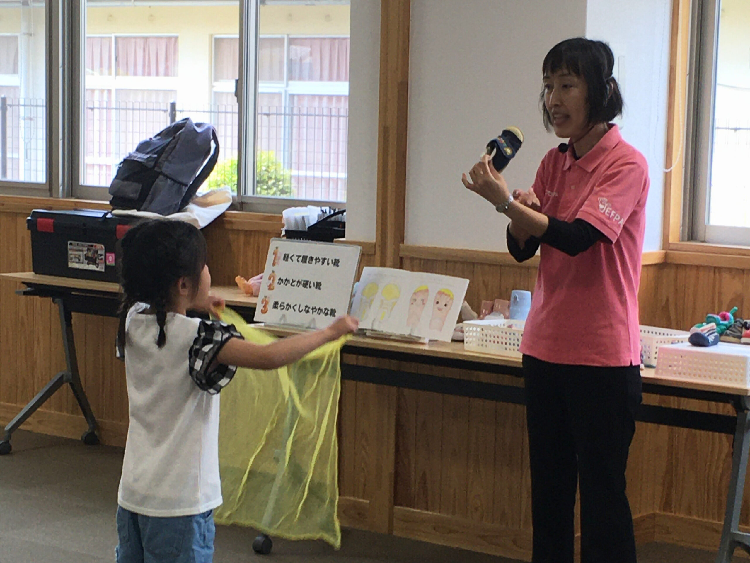 足育アドバイザーの成田が子ども用の靴を手に持ち、靴の話をしている様子。子どもが近くに寄ってきて、興味深そうに話を聞いている。