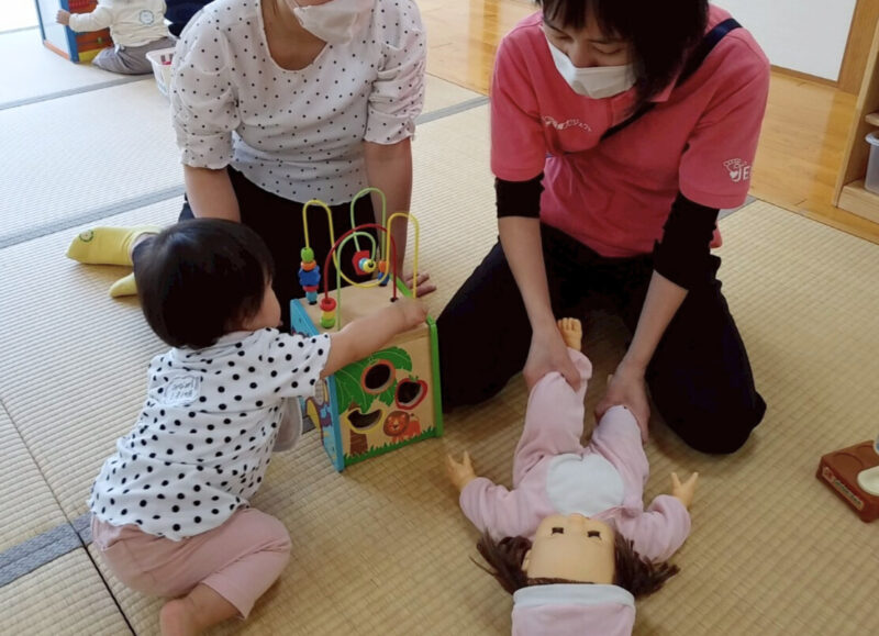 床に座って遊ぶ赤ちゃんを見守りながら、運動発達の相談をする保護者。講師の成田が隣で赤ちゃん人形を使って股関節の滑らかな動きを助ける遊びを実演して教えている様子(2025年5月8日、かのう子育て交流ひろば)