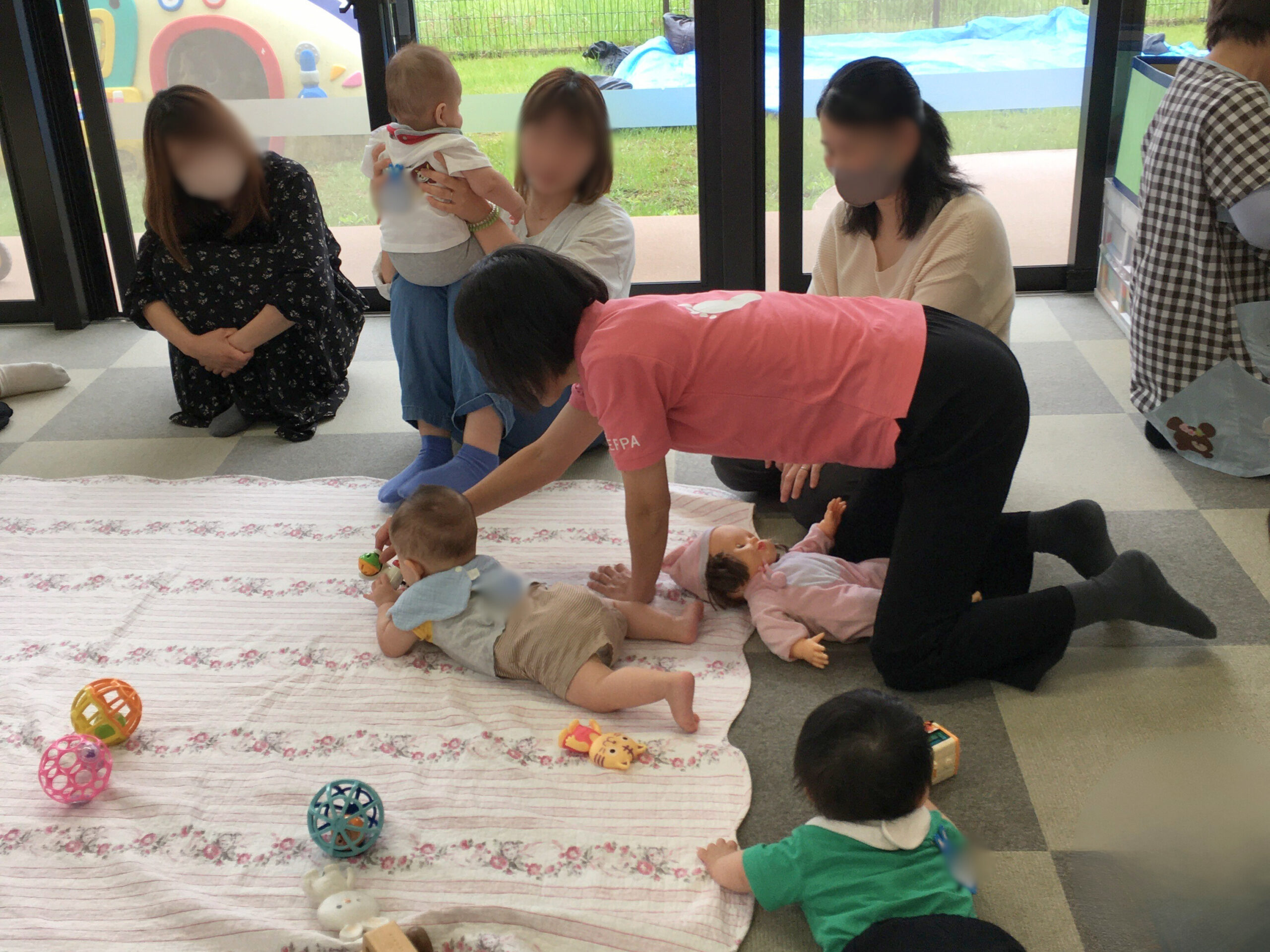 腹ばいを促すため、うつぶせの赤ちゃんの前におもちゃを置いて誘いかける講師。その実演を見守る母親たちの様子。(2025年5月29日)