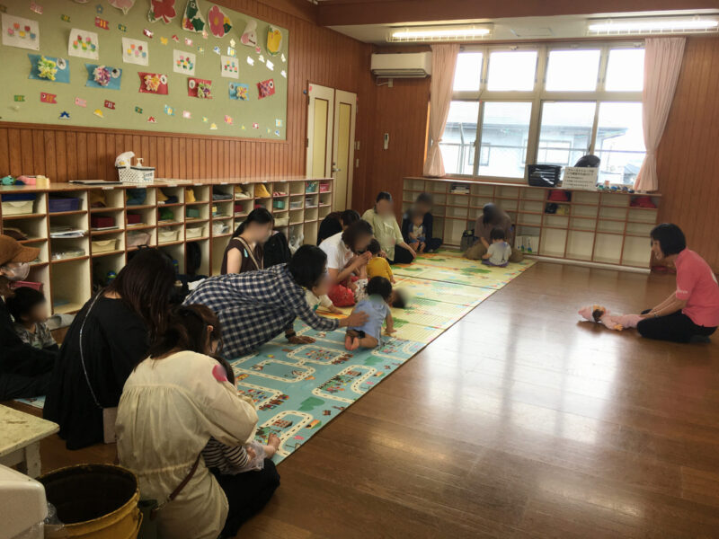 平和が丘幼稚園の子育て支援「すくすくクラブ」の講座風景。講師の成田の声かけにこたえて、マットの上でお母さんがわが子にふれる遊びをしている様子。(2025年5月21日)