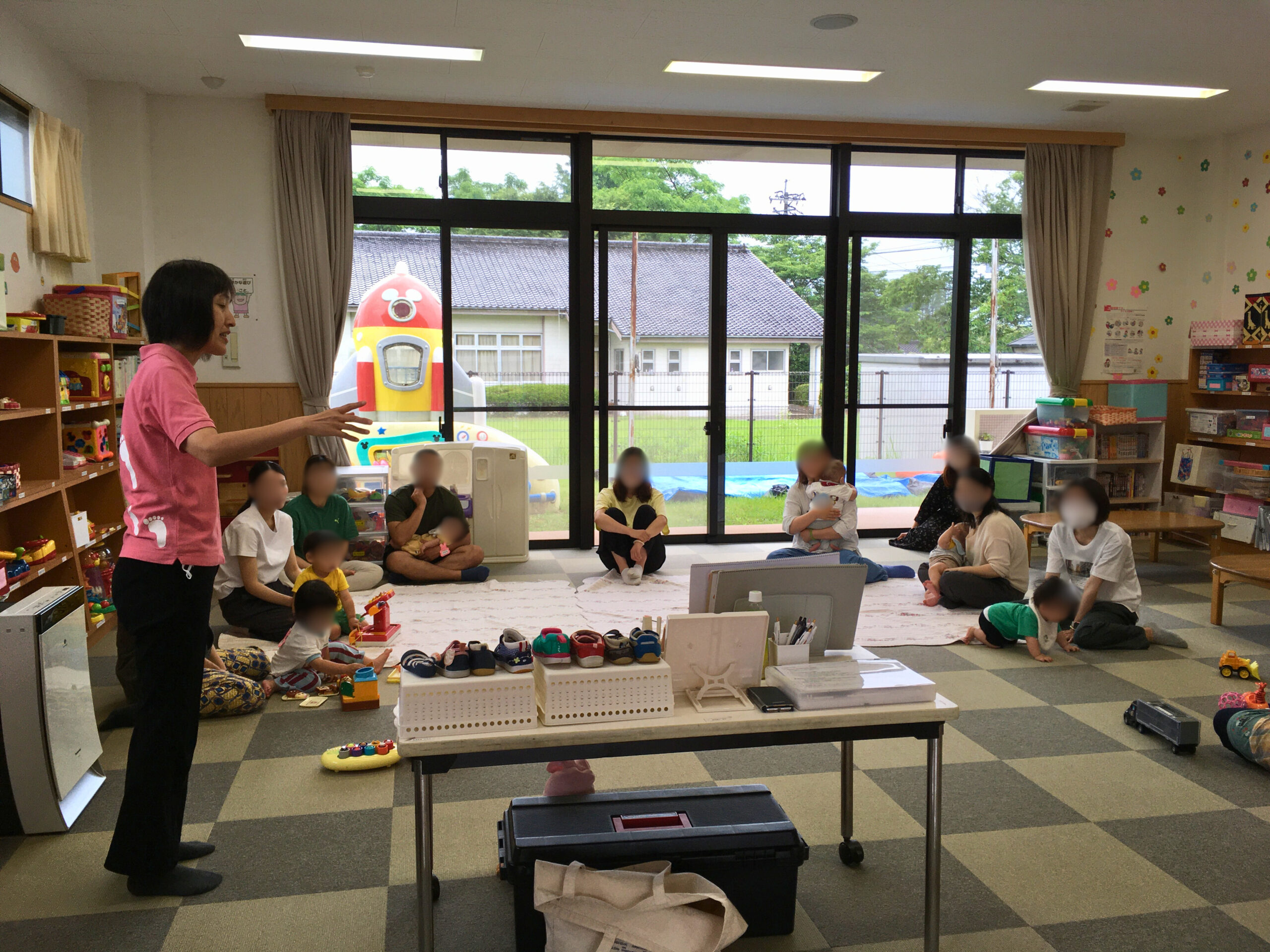 宮崎県国富町の森永児童館子育て支援センターで開催された足育講座。講師の成田が靴の選び方を説明しており、保護者は熱心に耳を傾けている。傍らでは乳幼児が遊び、お父さんの姿も見られる。(2025年5月29日)