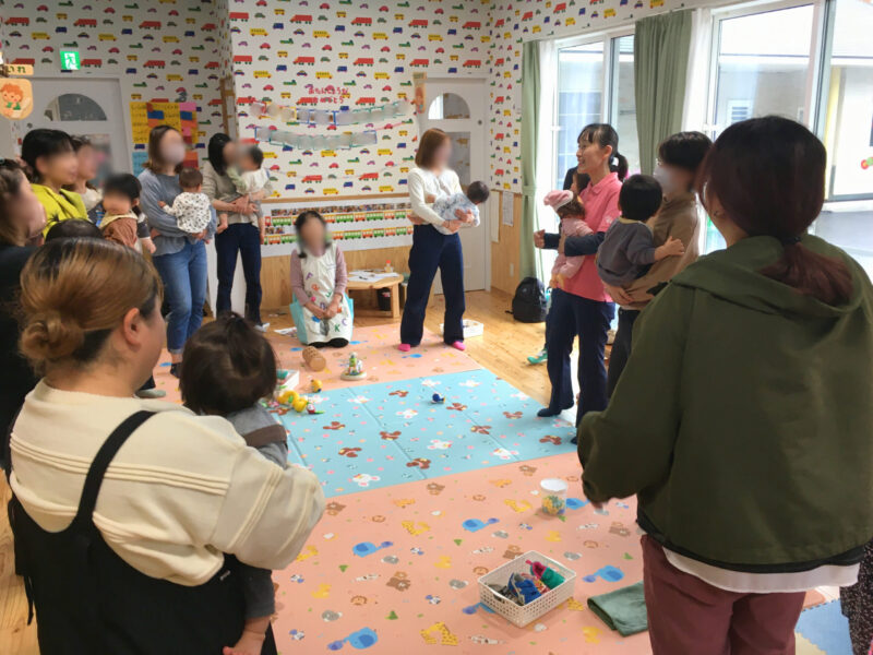 足育講座の様子。講師の説明を聞きながら、保護者が子どもを抱いて立ち、体をゆらす遊びを行っている。(2026年4月10日)