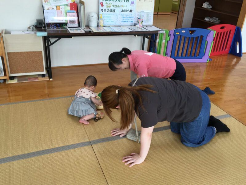 かのう子育て交流ひろばでの足育相談の様子。畳の上で、保護者がハイハイの姿勢を体験し、講師が体の使い方の違いを説明している。