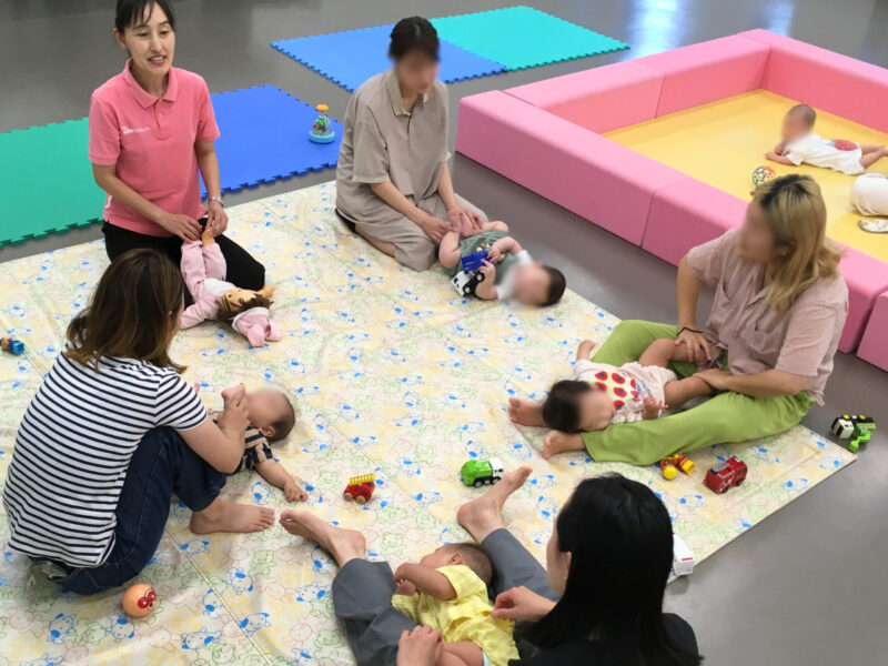お母さんと赤ちゃんが輪になって集まり、講師と一緒に赤ちゃんの足の運動遊びをしている様子